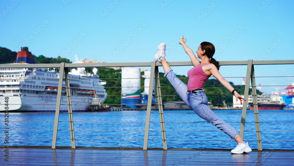 beautiful girl in life style denim clothes does twine on the railing in the port. dancer, gymnast, acrobat on sunny day in passenger port on background of cruise ship. twine on the observation deck beautiful girl in life style denim clothes does twine on the railing in the port. dancer, gymnast, acrobat on sunny day in passenger port on background of cruise ship. twine on the observation deck
