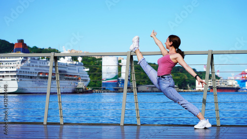 beautiful girl in life style denim clothes does twine on the railing in the port. dancer, gymnast, acrobat on sunny day in passenger port on background of cruise ship. twine on the observation deck