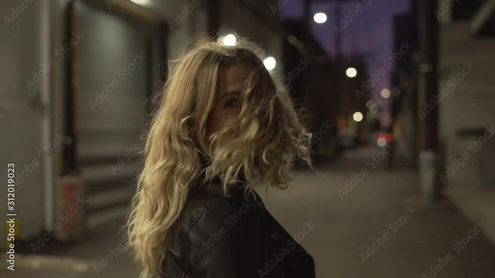 Slow motion of young blonde woman in jeans and leather jacket posing and walking on streets in downtown after sunset in the evening Slow motion of young blonde woman in jeans and leather jacket posing and walking on streets in downtown after sunset in the evening