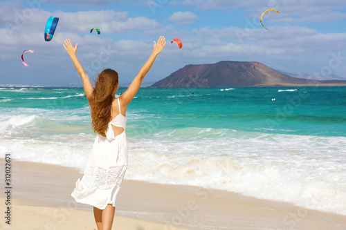 Fotografia Young woman enjoying wind with raised arms and people kitesurfering, Corralejo D
