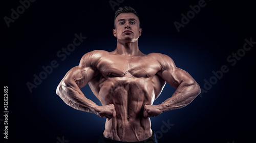 Handsome muscular bodybuilder posing on Front Lat Spread display lat width from the front, chest thickness, shoulder width, front arm and forearm size, quadriceps mass and separation