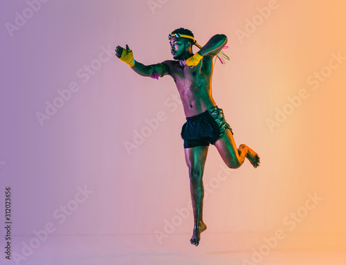 Muay thai. Young man exercising thai boxing on gradient background in neon light. Fighter practicing, training in martial arts in action, motion. Healthy lifestyle, sport, asian culture concept. © master1305
