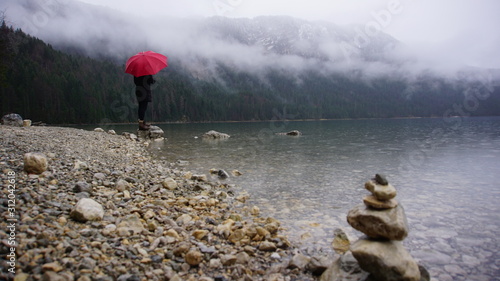 Wanderung um den Eibsee am Fuß der Zugspitze. Umgeben von grünen Tannen, Steinstrand und Felsbrocken. Nebel in den Bäumen. Leichter Regen. Wanderer mit rotem Regenschirm.