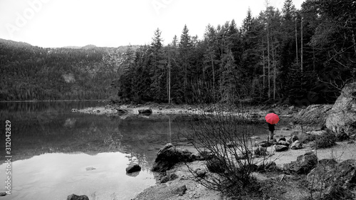 Walk around the Eibsea (Eibsee) near Zugspitze (highest mountain in Germany). Clear weather, no wind. Sea surrounded with green fir trees, grey stones and rock fragments. Wanderer with red umbrella.