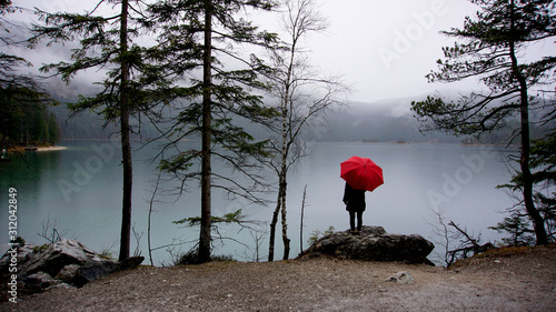 Walk around the Eibsea (Eibsee) near Zugspitze (highest mountain in Germany). Sea surrounded with green fir trees, grey stones and rock fragments. Foggy weather. Wanderer with red umbrella. Stone Art.