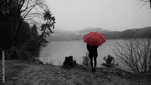 Walk around the Eibsea (Eibsee) near Zugspitze (highest mountain in Germany). Rainy weather. Sea surrounded with fir trees, stones and rock fragments. Wanderer with red umbrella. Black and white.