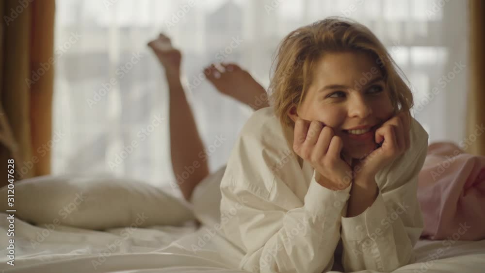Portrait of smiling Caucasian girl lying on soft bed and looking up. Positive young woman in white shirt shaking legs. Lifestyle, happiness, leisure. Portrait of smiling Caucasian girl lying on soft bed and looking up. Positive young woman in white shirt shaking legs. Lifestyle, happiness, leisure.