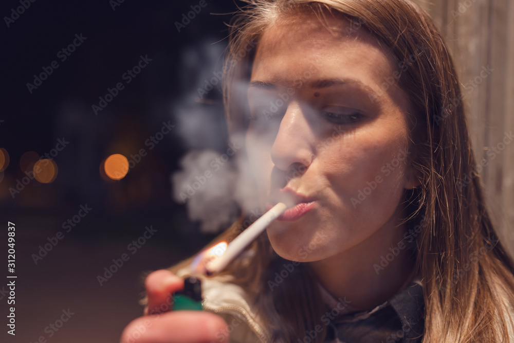 Fototapeta premium Close up portrait of young caucasian woman on the street at night lighting cigarette with lighter smoking in the city outdoor smoke
