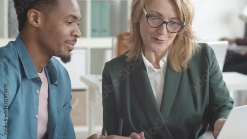 Tilting chest-up shot of middle-aged Caucasian female supervisor sitting at desk with laptop and explaining duties to young Afro-American intern on his first day of practice in office photo