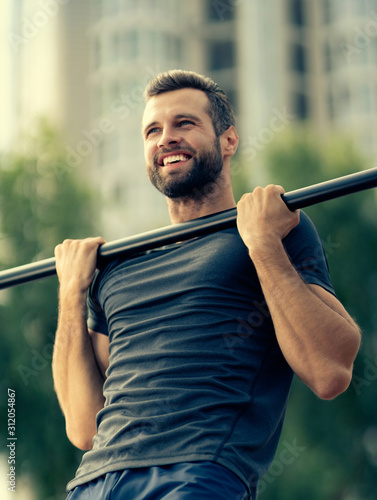 Portrait of happy bearded handsome man, doing pulling up exercise at horizontal bar, during workout training, outdoors. Fitness, sport, crossfit and workout in city concept.