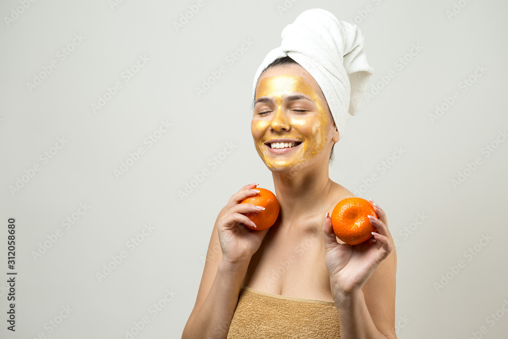 Beauty portrait of woman in white towel on head with gold nourishing mask on face. Skincare cleansing eco organic cosmetic spa relax concept. A girl stands with her back holding an orange mandarin. Beauty portrait of woman in white towel on head with gold nourishing mask on face. Skincare cleansing eco organic cosmetic spa relax concept. A girl stands with her back holding an orange mandarin.