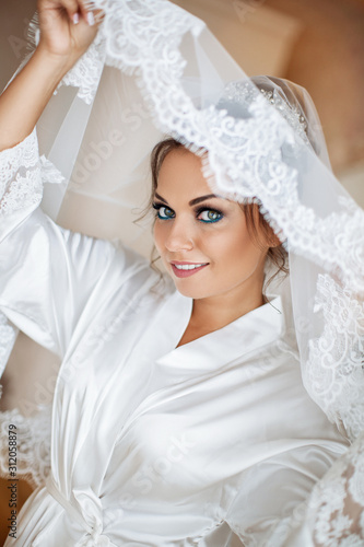 Happy beautiful bride under veil closeup. Beautiful bride lowered her eyes down, shows makeup and hairstyle. Bride in a white robe plays with her veil. Fashion, beauty, style. Morning of the bride. 