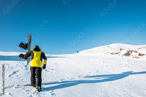 back view of sportsman holding snowboard while walking on white snow
