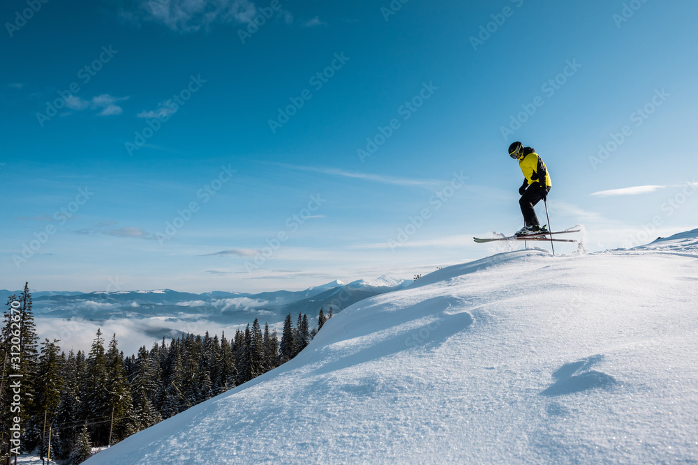 side view of skier holding ski sticks and jumping against blue sky in mountains