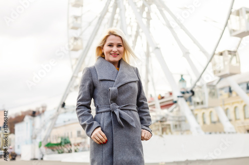 Stylish lady walking around city. Outdoor photo of pretty girl with charming smile posing in gray coat on urban background ferris wheel