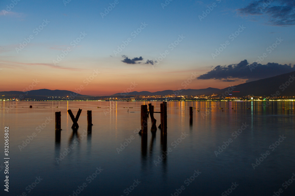 Evening city, the sea is beautifully reflected city lights. Mountains and sunset in the background. In the foreground, rusty crooked piles protrude from the water.