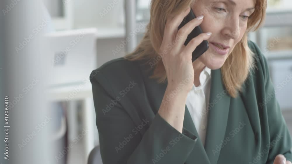 Tilting close-up shot of middle-aged Caucasian businesswoman in jacket sitting in office and having difficult conversation on mobile phone while looking at laptop screen