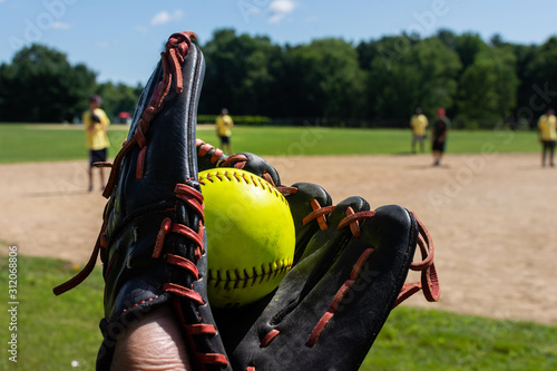 a man holds a freshly caught green softball in a black baseball glove during a softball tournament game with teammates in the background photo