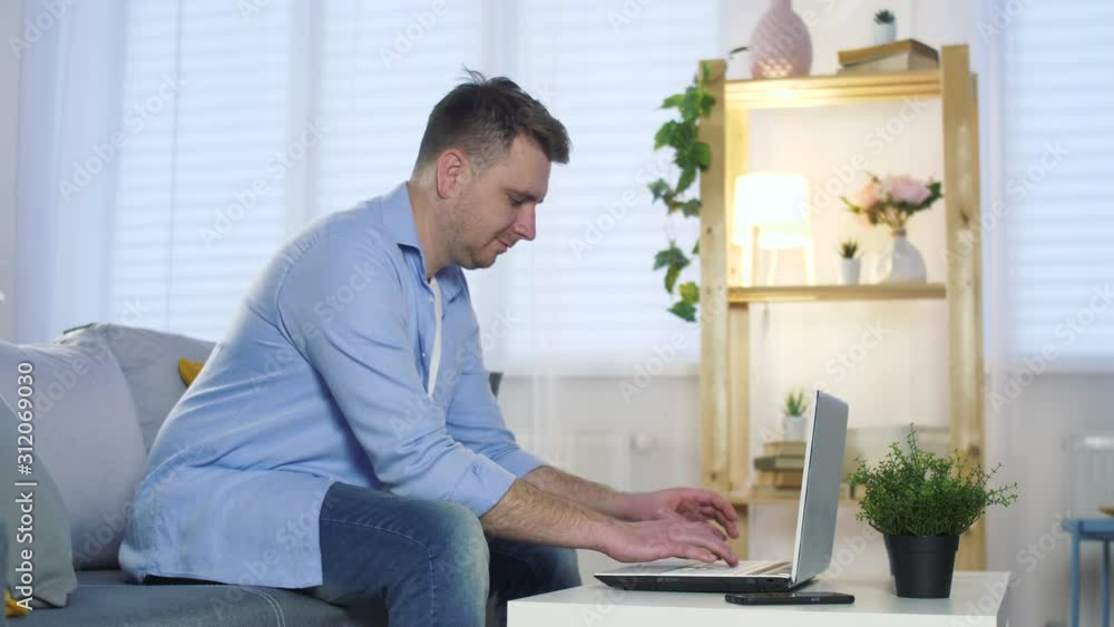 Young man using laptop for work or watching video or use social media. Boy seat on couch in modern living room. Relaxing at home. Slow motion. Young man using laptop for work or watching video or use social media. Boy seat on couch in modern living room. Relaxing at home. Slow motion.