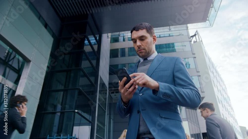 Wallpaper Mural Caucasian Businessman in a Suit is Using a Smartphone on a Street in Downtown. Other Office People Walk Past. He Smiles and Looks Successful. He's Browsing the Web on his Device. Torontodigital.ca