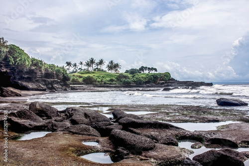 Tropical scenery in Tanah Lot beach with rocks, the ocean and palm trees in the background, during a cloudy day in Bali