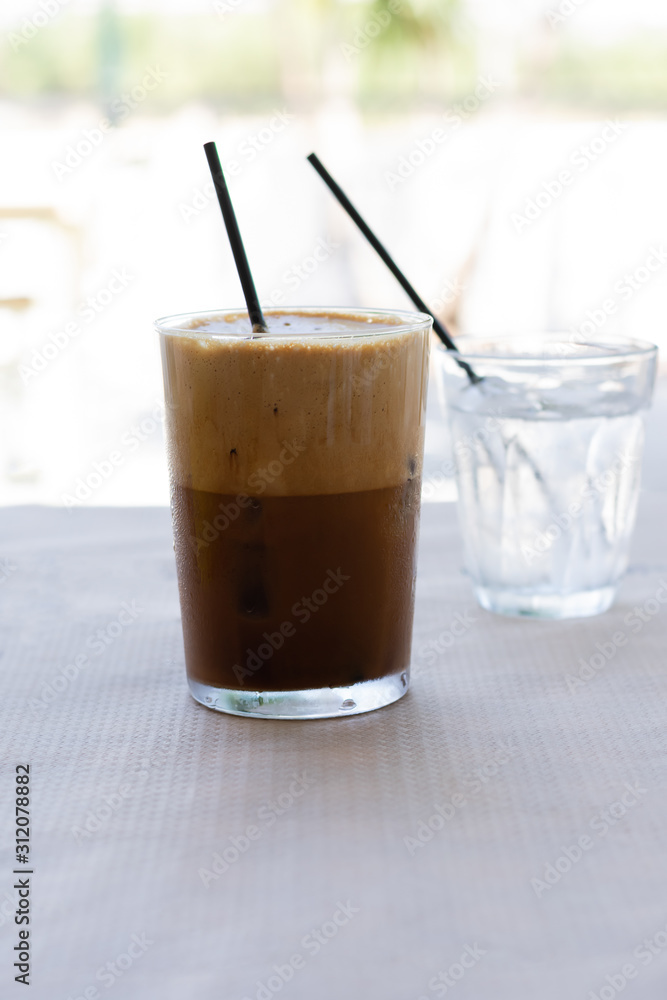 Cold frappe coffee with ice cubes on glass with black straw served on the table in caffeteria. Ice coffee in a tall glass with a lot of foam as cold summer drink. Cold frappe coffee with ice cubes on glass with black straw served on the table in caffeteria. Ice coffee in a tall glass with a lot of foam as cold summer drink.
