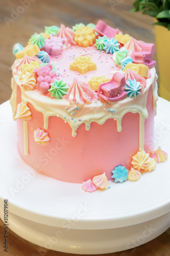 Party cake covered by pink and white cream with colorful meringue  chocolate bears  flowers and stars  and tiny confetti. Birthday party cake on white plate on wooden table. Vertical format.