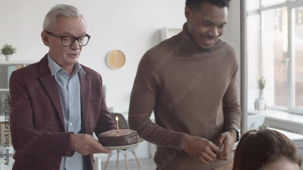 Medium tracking shot of two diverse male colleagues tiptoeing up to young Caucasian woman, congratulating her with birthday cake, delighted girl blowing out burning candle, all clapping and smiling Medium tracking shot of two diverse male colleagues tiptoeing up to young Caucasian woman, congratulating her with birthday cake, delighted girl blowing out burning candle, all clapping and smiling