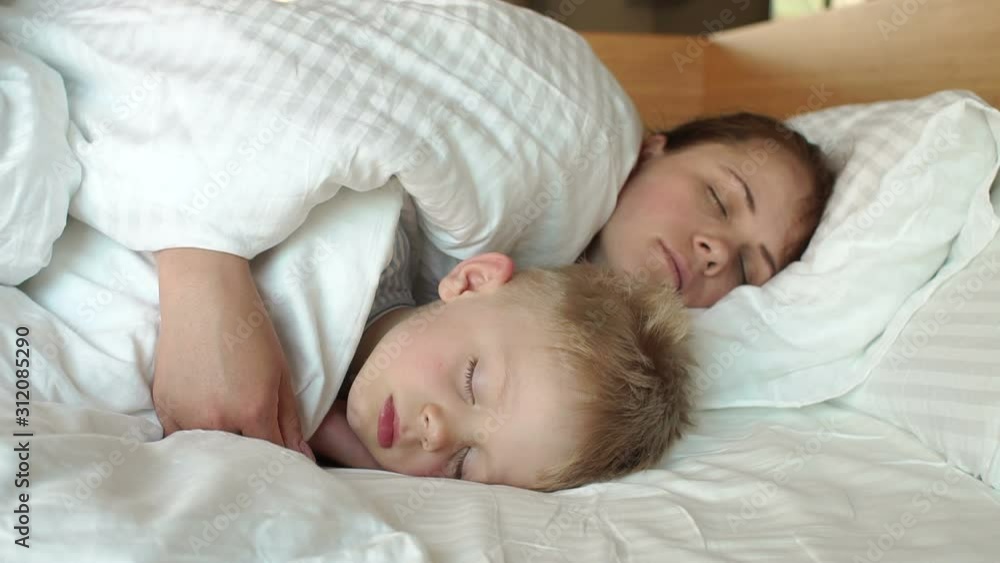 Close-up of mom gently hugging her sleeping son in the morning in bed. Mother and child sleep together in a white bed. Slow motion. Close-up of mom gently hugging her sleeping son in the morning in bed. Mother and child sleep together in a white bed. Slow motion.