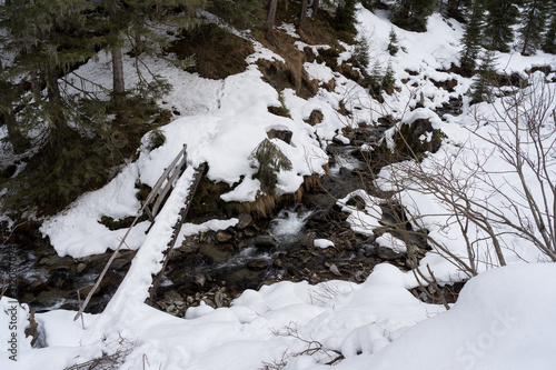 sehr schmale Brücke mit Schnee führt über einen Gebirgsbach