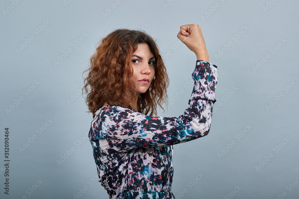 Serious charming young lady over isolated gray background in fashion shirt staying sideways, keeping fist up, looking at camera. People emotions. Lifestyle concept Serious charming young lady over isolated gray background in fashion shirt staying sideways, keeping fist up, looking at camera. People emotions. Lifestyle concept