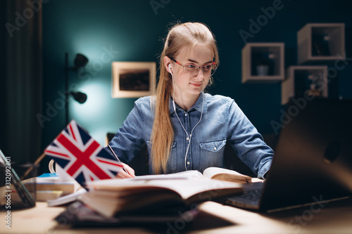 Concentrated girl in glasses and earphones studying English by a laptop at a desk