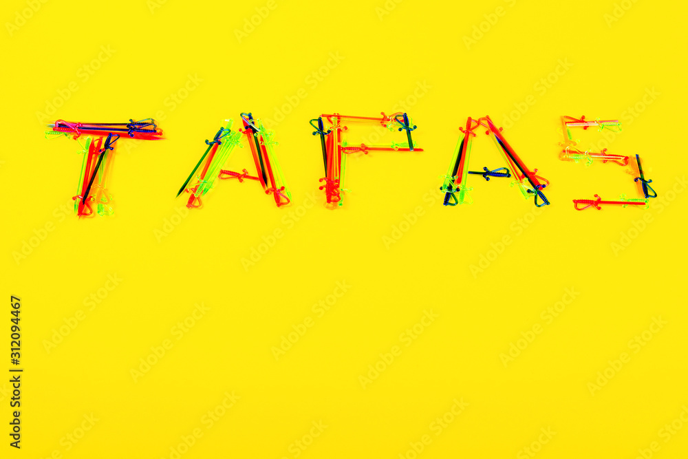 On yellow background is inscription Tapas, which is made of many multi-colored cocktail skewers. Such skewers are used to hold ingredients together in preparation of tapas. Copy space.