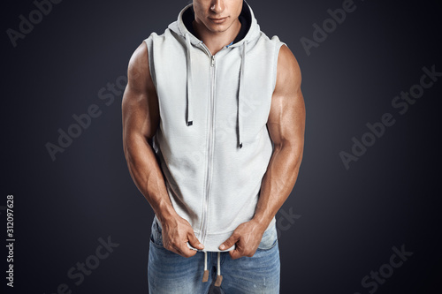 Close up photo of attractive fitness man wearing blank light gray sleeveless hoodie. Studio shot on dark background. Perfect fit, abdominal muscle, shoulders, deltoids, biceps. photo