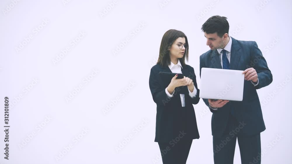 Pretty businesswoman standing with tablet in her hands, explaining something to her partner. Businessman on her side is holding laptop. They are checking data. Pretty businesswoman standing with tablet in her hands, explaining something to her partner. Businessman on her side is holding laptop. They are checking data.