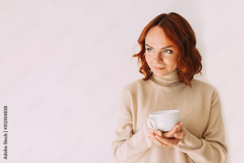Lifestyle image of red haired beautiful girl sipping coffee from a white mug, wearing cozy sweater of a pastel color. Looking aside. Copy space. Lifestyle image of red haired beautiful girl sipping coffee from a white mug, wearing cozy sweater of a pastel color. Looking aside. Copy space.