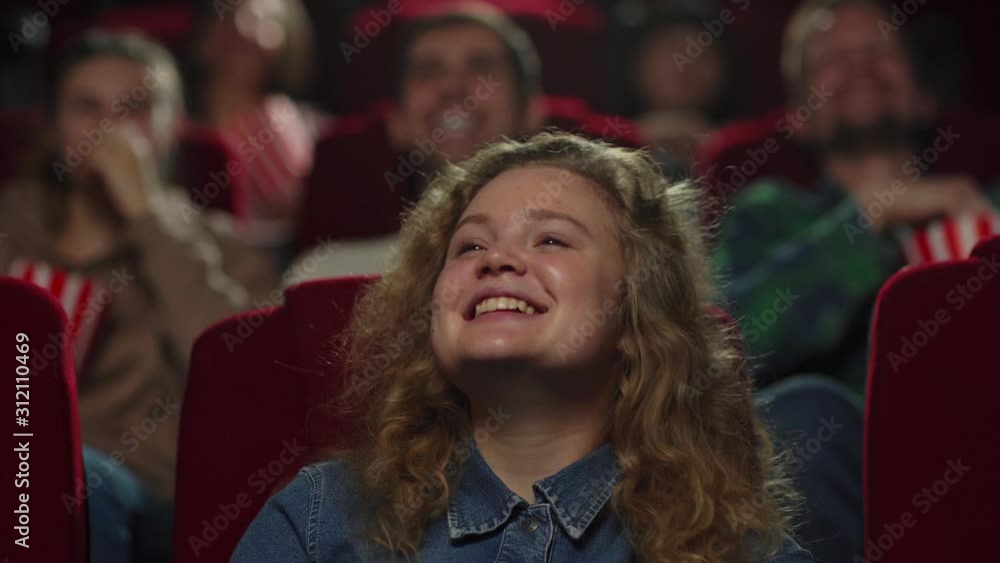 Zoom in closeup shot of curly-haired young woman laughing joyfully while watching comedy film in movie theater Zoom in closeup shot of curly-haired young woman laughing joyfully while watching comedy film in movie theater