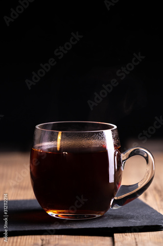  a transparent glass cup with hot tea stands on an old wooden surface on a black background with a place for inscription