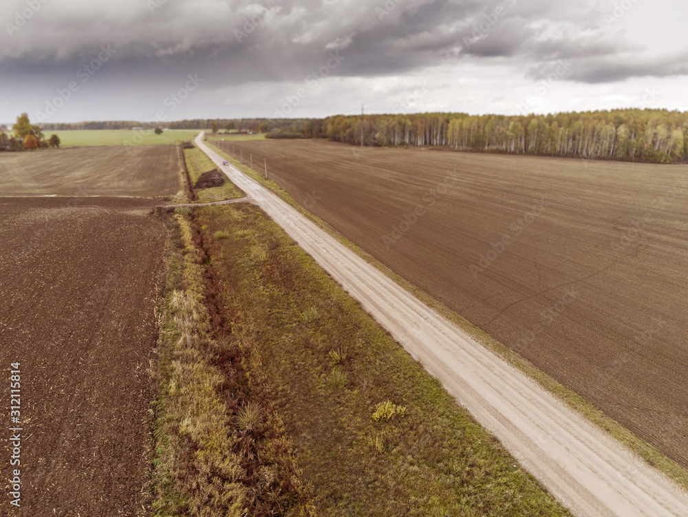 Small road in a country side by ploughed agricultural fields. Cloudy sky, Selective focus, Forest in the background. Aerial  view.