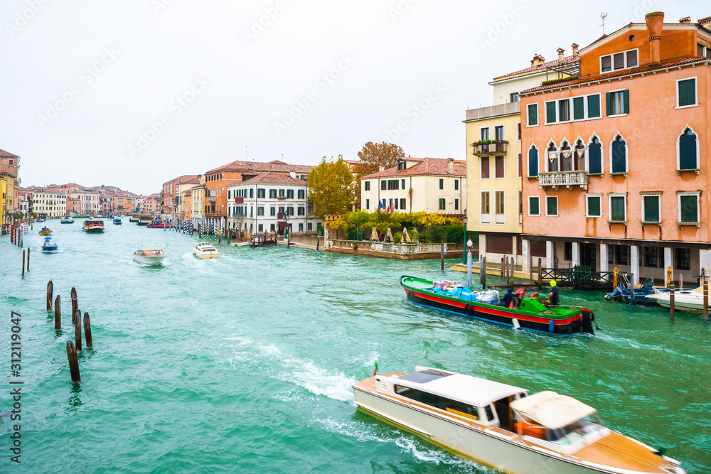 Fototapeta premium Boats, yachts, vaporettos, water taxis sailing down the Grand Canal waterway between wooden and striped mooring poles and colorful Venetian architecture buildings. November in Venice city, Italy.