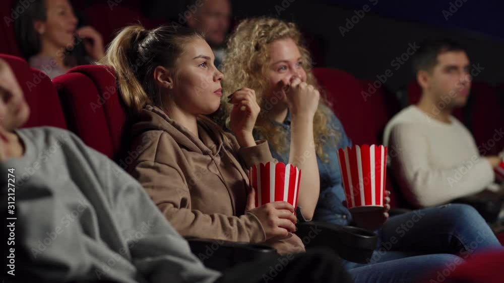 Side view medium shot of two cheerful young women eating popcorn and talking while watching movie sitting on comfortable seats in cinema Side view medium shot of two cheerful young women eating popcorn and talking while watching movie sitting on comfortable seats in cinema