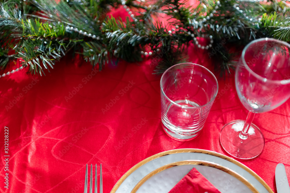 Table setup lunch in red for celebration. Shiny glass cup, red tablecloth, candles and decorations with green Christmas tree. Christmas dinner during the holiday decorated table. Dining table setup Table setup lunch in red for celebration. Shiny glass cup, red tablecloth, candles and decorations with green Christmas tree. Christmas dinner during the holiday decorated table. Dining table setup