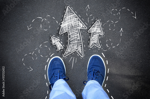 Person standing on asphalt road near arrows pointing in different directions, top view. Concept of choice