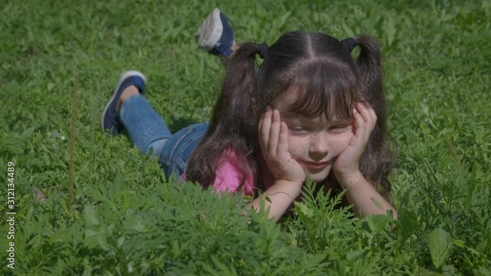 The child lies on a green lawn. A cute little girl is lying on the grass and swinging her legs in pleasure. The child lies on a green lawn. A cute little girl is lying on the grass and swinging her legs in pleasure.