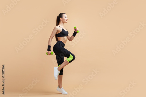 A young brunette girl raises her knees high, using extra weights, a full-length photo, against a peach background.