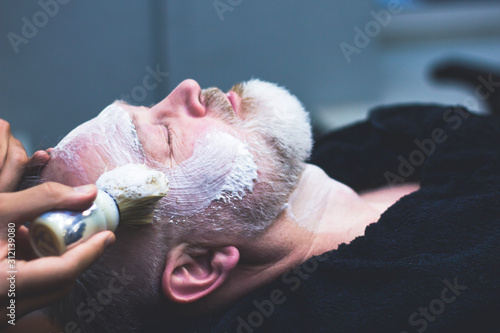 An older man is shaved in a barber shop. a professional worker applies shaving foam to the face of a client. Beauty and care for your appearance