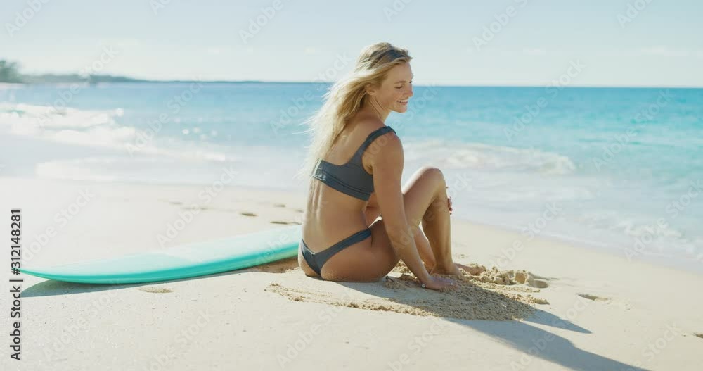 Beautiful young woman in a bikini sitting on a sunny beach smiling and enjoying her tropical vacation, attractive blonde surfer sitting on the beach with her blue surfboard Beautiful young woman in a bikini sitting on a sunny beach smiling and enjoying her tropical vacation, attractive blonde surfer sitting on the beach with her blue surfboard