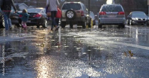 A low angle shot of the legs of a cold family wearing raincoats quickly walking to their cars on a rainy evening as fall leaves get soaked on the pavement.