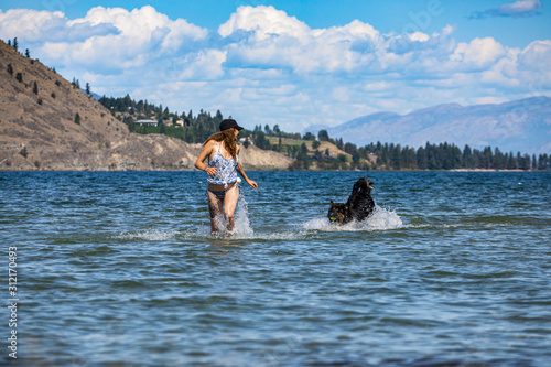 close up of cute girl with swimsuit and black hat plays on the sand and inside the crystal water of the river with her dog in british columbia... © Valmedia