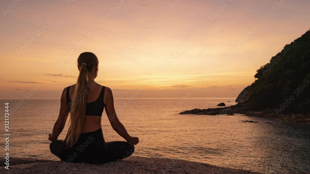 Young woman sitting and meditating in lotus position on a rock by the sea against the background of sunrise 4k time lapse Young woman sitting and meditating in lotus position on a rock by the sea against the background of sunrise 4k time lapse
